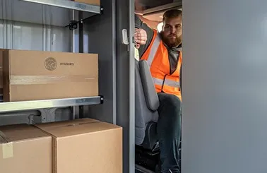 a man wearing a safety vest sitting in the driver's seat of a van, opening a sliding partition that leads to the cargo area