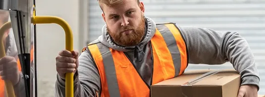 man wearing a safety vest holding a box as he enters a work van with the aid of a grab handle by Adrian Steel