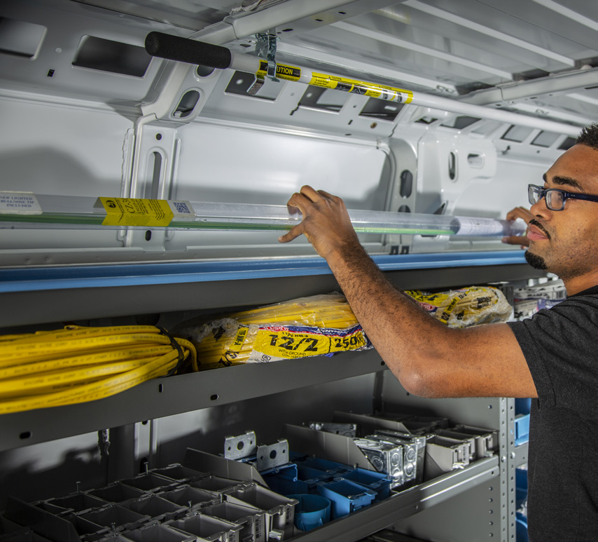 a man in a fully upfitted van placing a long object on an Adrian Steel long part storage solution